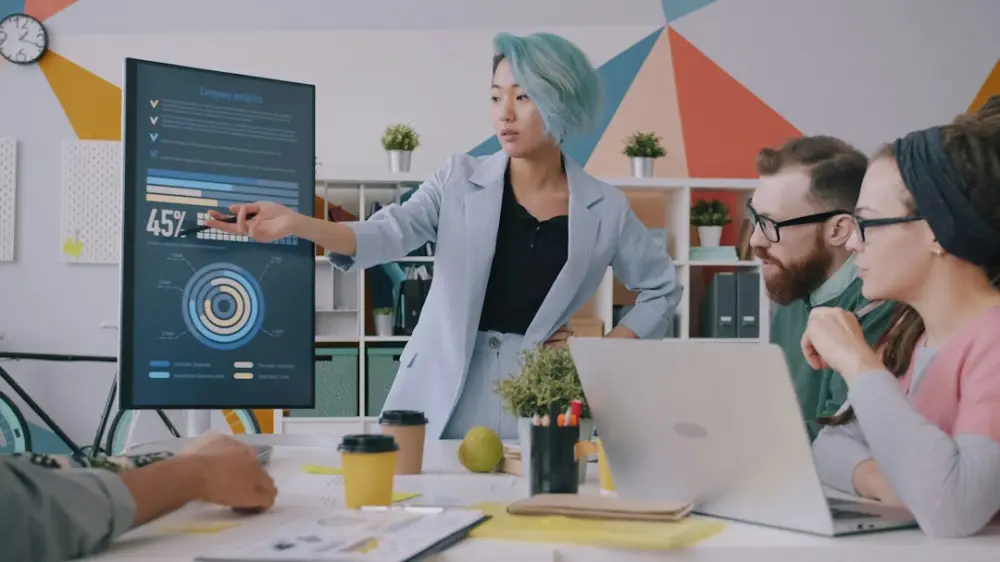 A woman with blue hair pointing at a digital chart during a team meeting, illustrating measurement and conversion